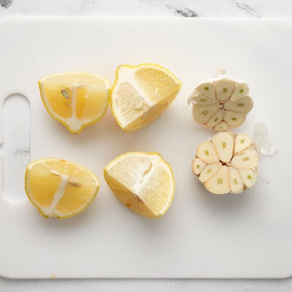 Des quartiers de citron et une gousse d'ail coupée en deux sur une planche à découper, en préparation pour un rôti de poulet.