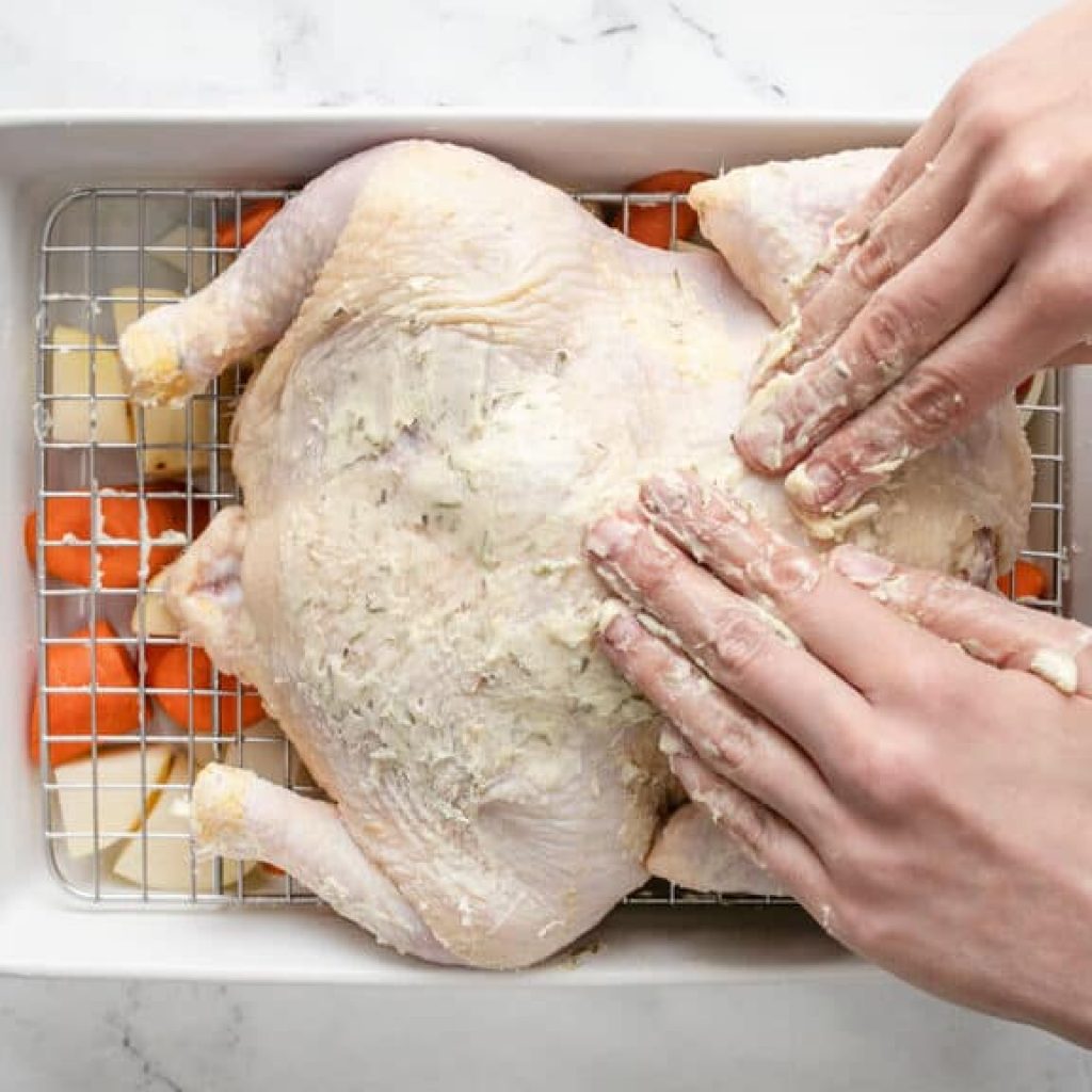Des mains frottent un mélange de beurre et de romarin sur un poulet cru dans un plat de cuisson avec des carottes et des pommes de terre, en préparation pour un rôti de poulet.