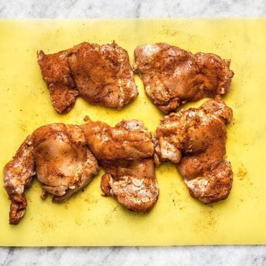 Hauts de cuisses de poulet crus, généreusement saupoudrés d'épices rouges et brunes, posés sur une planche jaune pour la préparation du Tikka Masala.