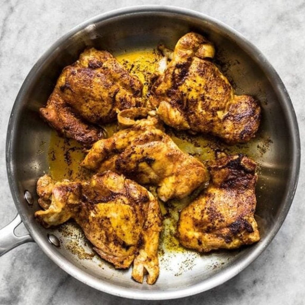 Gros plan de hauts de cuisses de poulet assaisonnés, qui sont en train de cuire et de caraméliser dans une poêle. On aperçoit un peu de matière grasse fondue. Préparation du Tikka Masala.