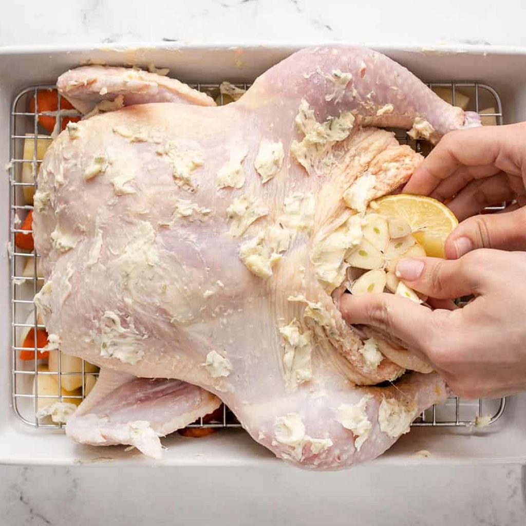 Des mains remplissent la cavité d'un poulet beurré avec des quartiers de citron et des gousses d'ail, en préparation pour un rôti de poulet.