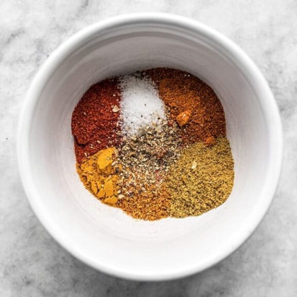 Assortiment d'épices non mélangées (paprika, sel, poivre, curcuma et cumin) dans un bol blanc, préparation pour du poulet tikka masala.