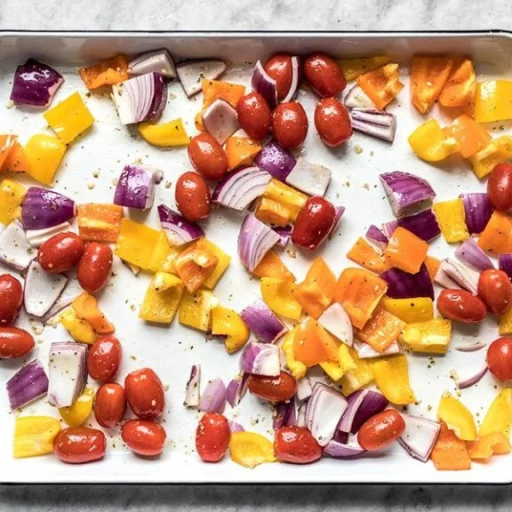 Une plaque de cuisson avec un assortiment coloré de légumes : tomates rouges, poivrons jaunes et orange, et oignons violets, qui accompagneront un poulet grec rôti au four.