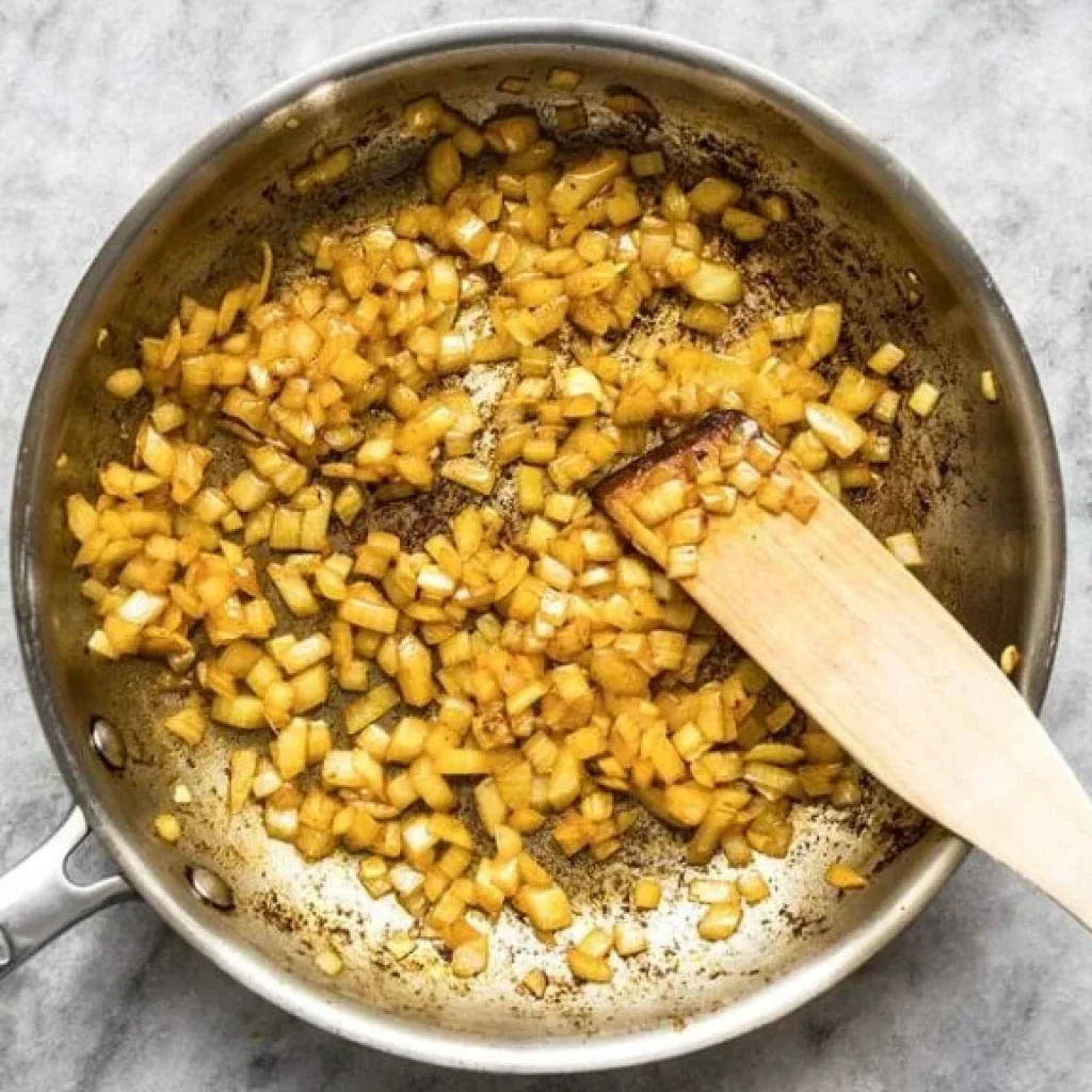 Photo en plongée d'oignons coupés en dés, cuisant dans une poêle en acier inoxydable avec une cuillère en bois. Les oignons commencent à caraméliser, une étape importante pour le Tikka Masala.