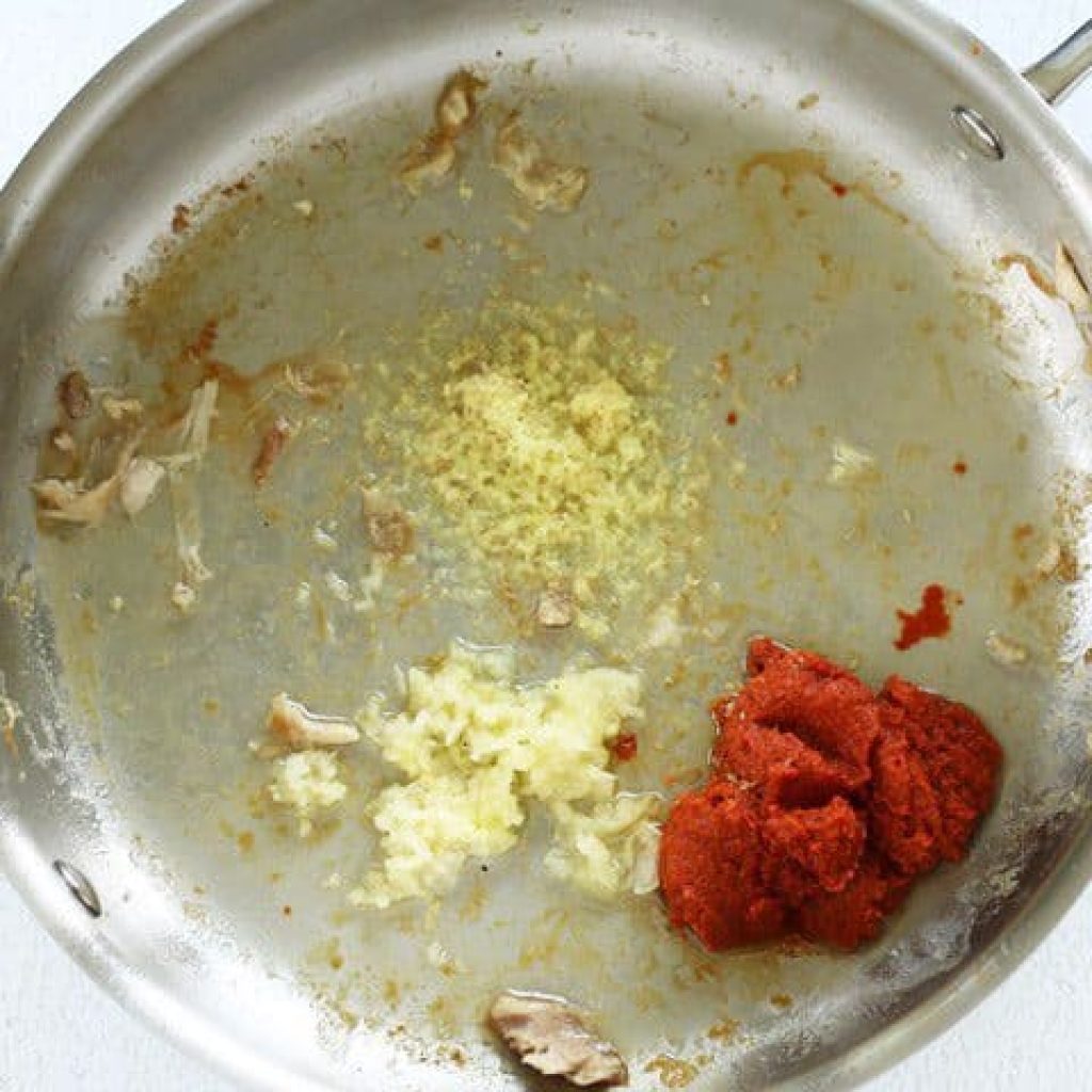 une poêle en acier inoxydable avec des restes de poulet, du gingembre râpé, de l'ail haché et de la pâte de curry rouge."