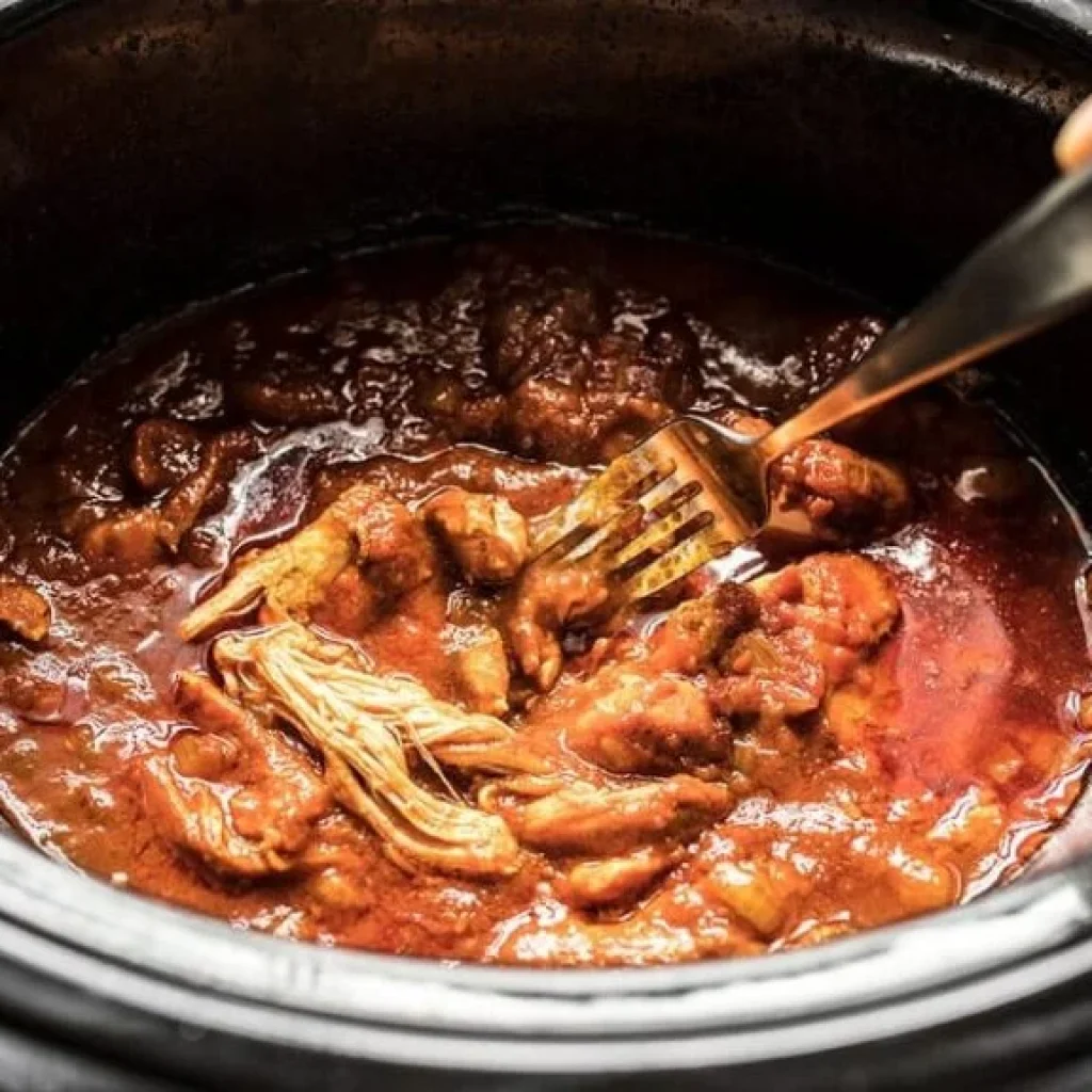 Poulet Tikka Masala effiloché en train de mijoter dans une mijoteuse. La sauce est épaisse et appétissante. On voit une fourchette qui mélange les ingrédients.