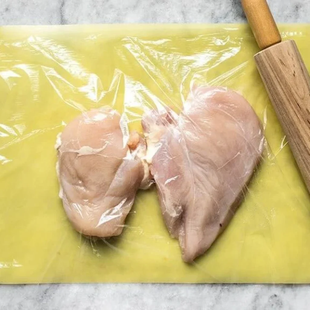 "Des poitrines de poulet crues, protégées par du film alimentaire, avant d'être préparées pour la cuisson d'un délicieux poulet grec au four."