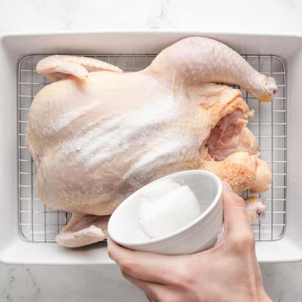 Une main verse du sel sur un poulet cru placé sur une grille dans un plat de cuisson, en préparation pour une recette de poulet rôti.