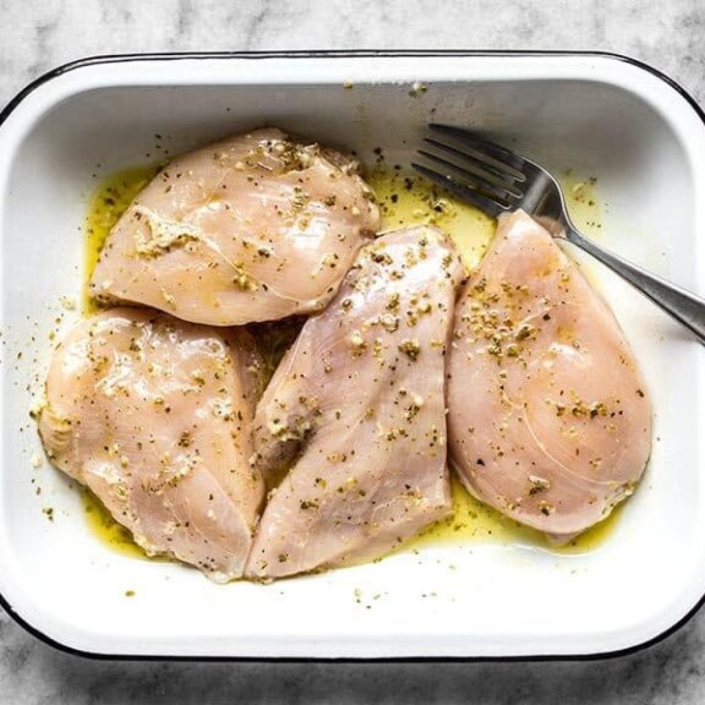 "Quatre poitrines de poulet crus, immergés dans une marinade à l'huile d'olive et aux épices, sur une plaque, en préparation pour un poulet grec rôti."
