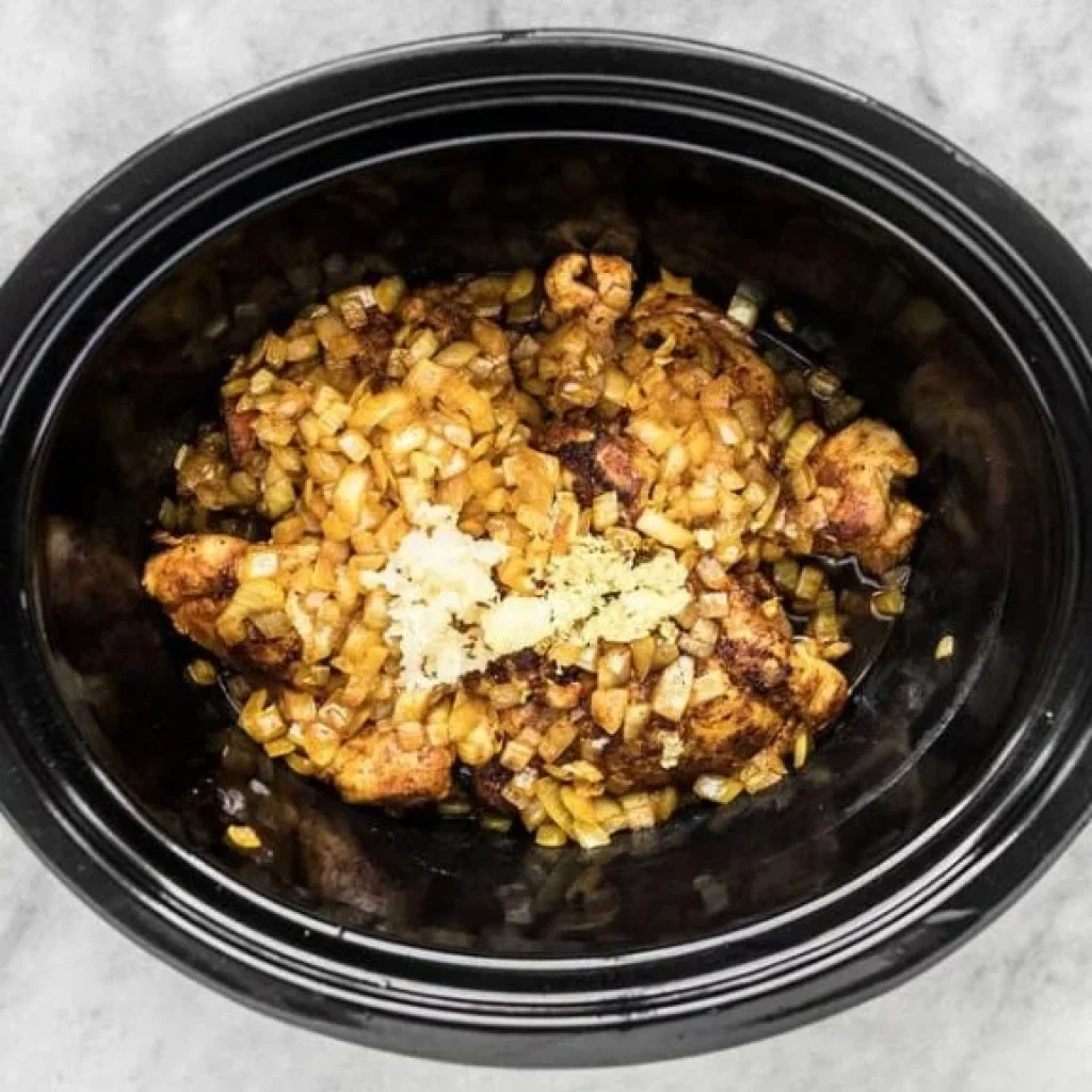 Dans une mijoteuse, on voit du poulet doré, des oignons cuits et de l'ail haché. Les ingrédients se mélangent pour la préparation d'un poulet Tikka Masala.