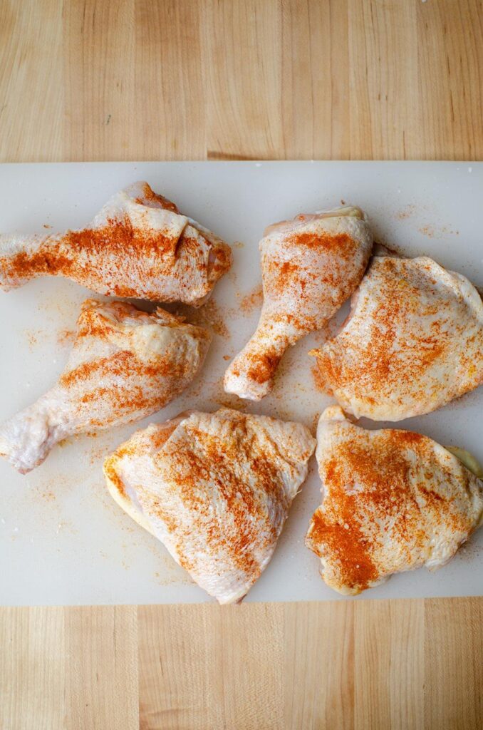 "Morceaux de poulet crus, saupoudrés d'épices rouges, disposés sur une planche à découper. Préparation pour un poulet basquaise à l'ancienne."