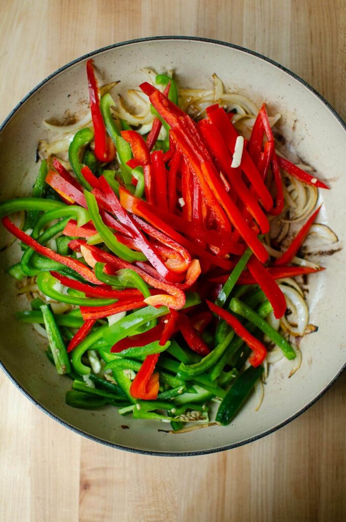 Poivrons rouges et verts coupés en lamelles, ajoutés aux oignons dorés dans une poêle. Étape de la préparation du poulet basquaise à l'ancienne.