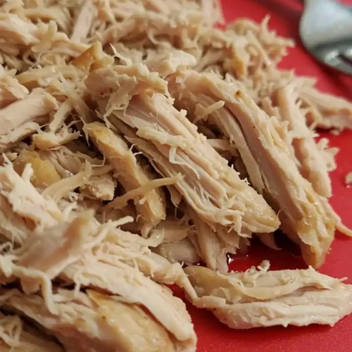 De la viande de poulet cuite et effilochée reposant sur une planche à découper rouge. Elle est prête à être ajoutée à une soupe au poulet et aux nouilles.