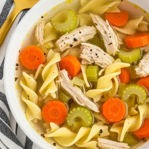 Un bol blanc rempli d'une soupe au poulet et aux nouilles. On peut voir des morceaux de poulet, des carottes, du céleri et des pâtes.