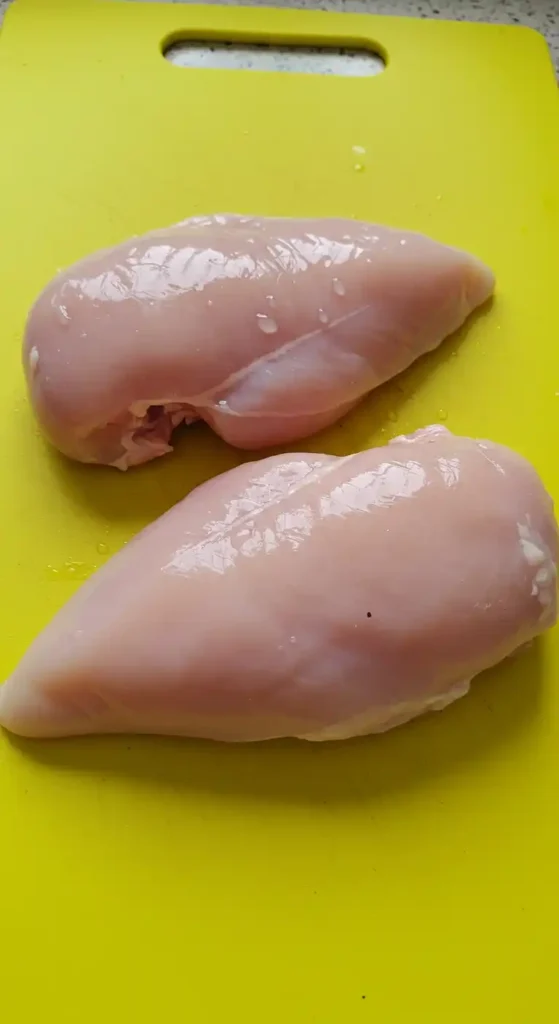Deux poitrines de poulet crues, posées sur une planche à découper jaune. Elles sont prêtes à être cuites pour une soupe au poulet et aux nouilles.