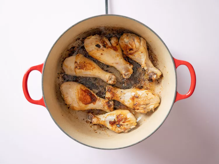 Une vue aérienne de plusieurs pilons de poulet en train de dorer dans une cocotte rouge sur une surface blanche.