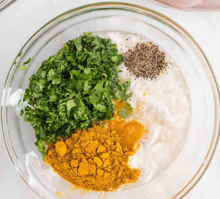 Bol en verre rempli d'ingrédients pour une marinade poulet curry : le blanc crémeux du yaourt contraste avec le vert vif de la coriandre fraîche hachée, le jaune chaud et poudré du curry et le noir du poivre moulu, promettant une marinade parfumée et savoureuse.

