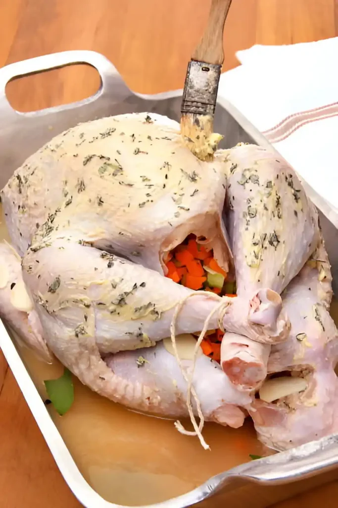 Un poulet entier préparé pour être rôti au four, badigeonné de beurre aux herbes, farci de légumes frais comme des carottes et du céleri, et placé dans un plat à rôtir en métal avec un peu de bouillon au fond.