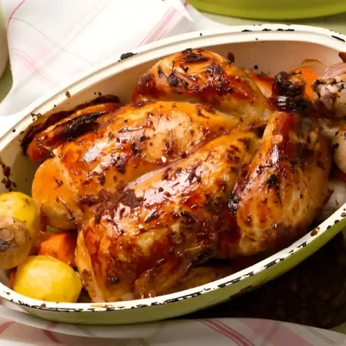 Un poulet rôti doré dans un plat, avec des pommes de terre, des carottes et des oignons caramélisés.