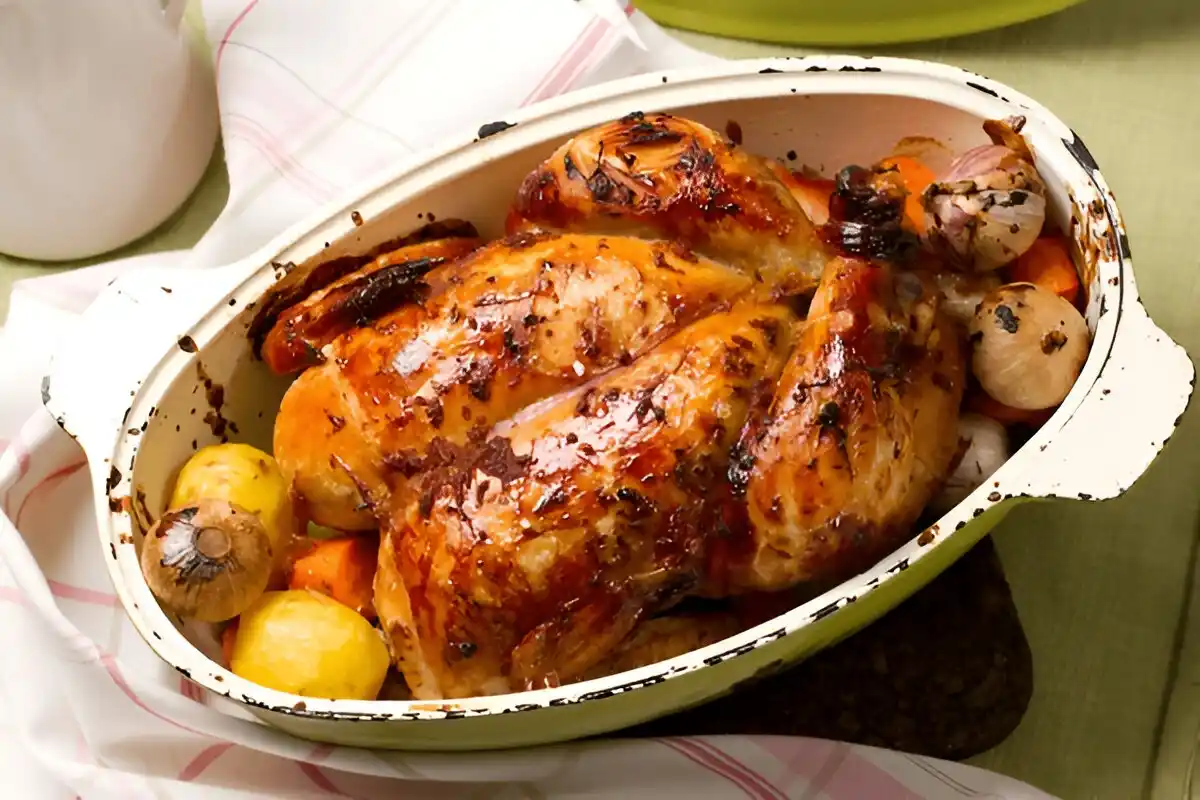Un poulet rôti doré dans un plat, avec des pommes de terre, des carottes et des oignons caramélisés.