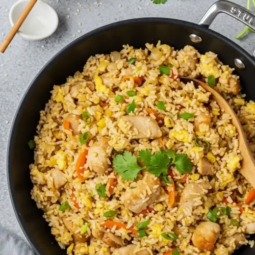 Image de riz frit au poulet dans une poêle noire, avec du poulet, des carottes, des œufs et des oignons verts. Des graines de sésame et de la coriandre fraîche sont visibles à côté de la poêle avec une cuillère en bois