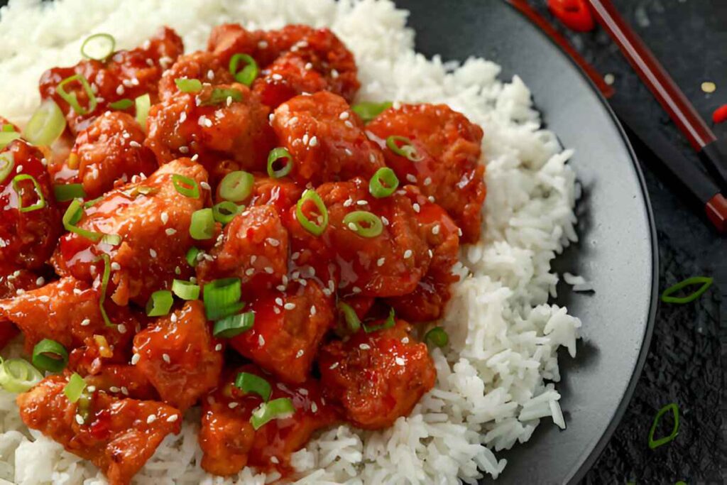 "Une prise de vue rapprochée, en plongée, de poulet Général Tao servi sur un lit de riz blanc. Les morceaux de poulet sont enrobés d'une sauce rouge brillante et sont garnis d'oignons verts émincés et de graines de sésame. L'assiette est noire, et une partie de baguettes de couleur foncée est visible sur le côté droit. Des morceaux d'oignon vert dispersés sont présents sur la surface texturée sombre