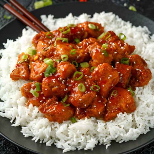 Poulet Général Tao croustillant nappé d'une sauce rouge brillante, servi sur un lit de riz blanc. Garniture de cébette et graines de sésame."
